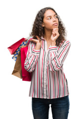 Young hispanic woman holding shopping bags serious face thinking about question, very confused idea