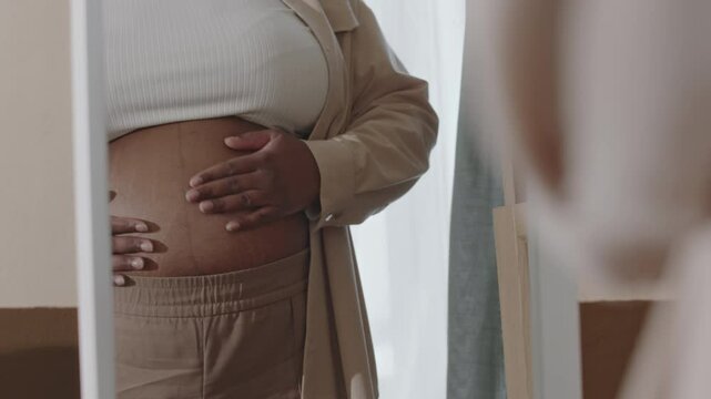 Mirror reflection of happy young pregnant African American woman looking at herself admiring changed appearence and rounded abdomen, caressing stomach with affection