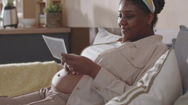 Medium tilt of pregnant young African American woman holding and admiring ultrasound image of future child while resting on comfortable couch at home