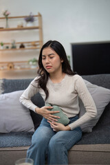 Obraz premium Young Woman Experiencing Abdominal Pain While Sitting on a Couch at Home, Holding a Hot Water Bottle for Relief