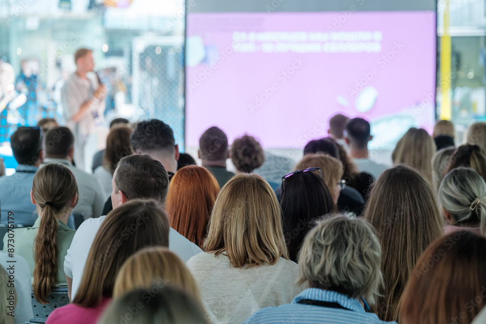 Wall mural A large audience sits attentively as a speaker delivers a presentation during a business seminar. The seminar takes place in a modern, well-lit hall.