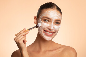 Happy young beautiful woman with bare shoulders applying white cosmetic clay on her face against...