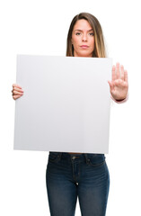 Fototapeta premium Beautiful young woman holding advertising banner with open hand doing stop sign with serious and confident expression, defense gesture