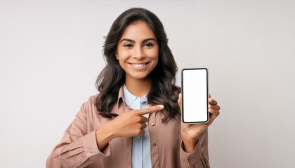 Naklejka premium A young modern happy Indian Asian woman or female is showing and pointing a finger at a mobile phone or smartphone blank screen and smiling looking at the camera isolated on a white background.