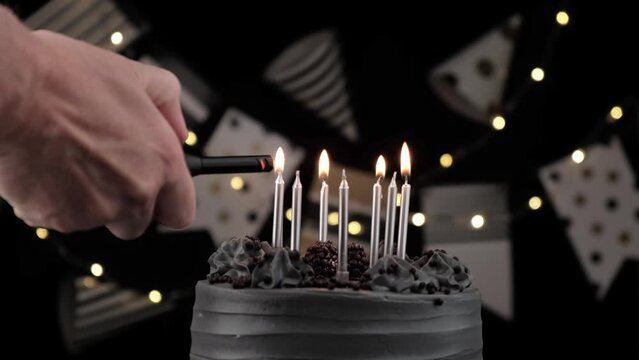man hand lights silver candles on black birthday cake on black background with garlands and flags. close-up bright candles on birthday cake. family traditions, fun holidays.