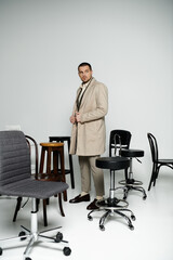 A confident man exudes timeless elegance in a room with varied chairs.