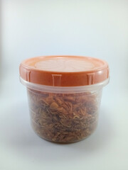 Deep-fried savory crispy sliced shallots in a transparent container with a cream-orange cover, placed against a white background, offering a crunchy texture and rich flavor in a visually simple place