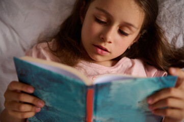 Relaxed little student girl lying on the bed at home, reading a fairy tale book while the story bedtime. People. Happy carefree childhood. Education and erudition. The concept for World Book Day
