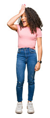 Young beautiful woman with curly hair wearing pink t-shirt surprised with hand on head for mistake,...