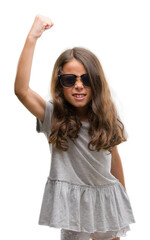 Brunette hispanic girl wearing sunglasses angry and mad raising fist frustrated and furious while shouting with anger. Rage and aggressive concept.