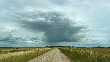 Panorama shot of a rural dirtroad in a flat US midwestern like landscape leading straight into a huge thunderstorm cloud hanging over the horizon