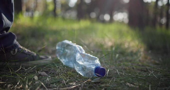 A man indifferently kicks a trash bottle lying on the ground in the forest without picking it up