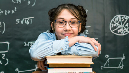 A schoolgirl girl put her hands and head on a stack of books in a school class.