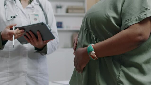 Medium shot of female Caucasian gynecologist standing in clinic and talking with African american woman expecting baby, scheduling next appointment using tablet