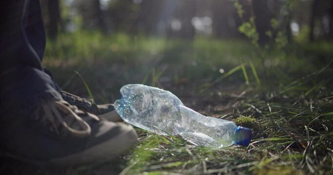A man indifferently kicks a trash bottle lying on the ground in the forest without picking it up