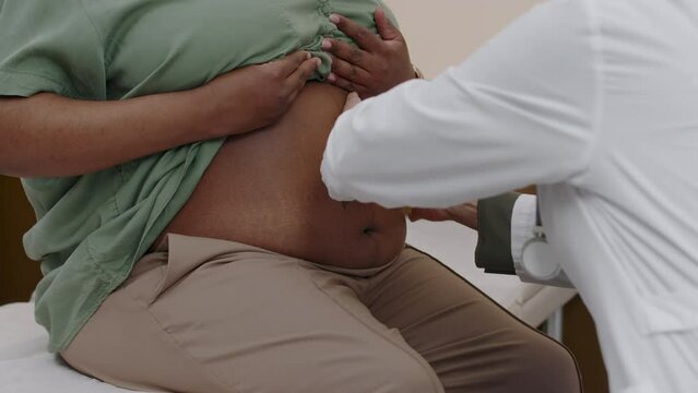 Unrecognizable female gynecologist doing physycal examination and checking position of unborn baby by touching abdomen of pregnant African American woman during scheduled visit