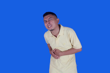 Funny face of Asian man wearing yellow t-shirt suffering from stomach ache due to diarrhea on blue...