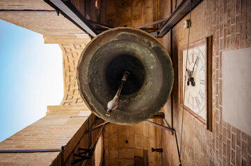 Bottom view of the inside of a large metal bell located inside a stone tower with an antique clock on the wall