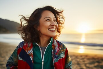 Portrait of a grinning asian woman in her 50s sporting a stylish varsity jacket in front of stunning sunset beach background