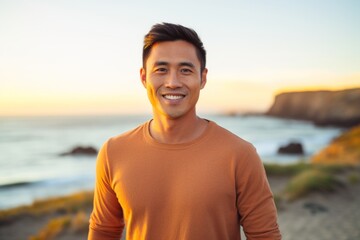 Portrait of a smiling asian man in his 30s showing off a thermal merino wool top on vibrant beach sunset background