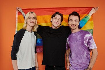 Three young friends stand together in front of an orange background, holding a rainbow flag and smiling.