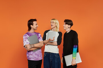 Three young queer friends in stylish attire stand against an orange background, laughing and holding laptops.