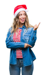 Beautiful young blonde woman wearing christmas hat over isolated background with a big smile on face, pointing with hand and finger to the side looking at the camera.