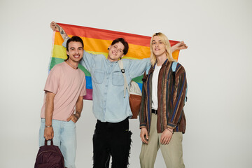 Three young queer students stand together, holding a rainbow flag in front of a grey background.