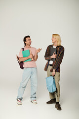 Fototapeta premium Two young adults, dressed in casual attire, stand against a grey background, talking and laughing.