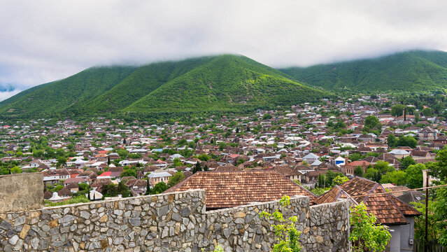 A view of the city of Shaki in Azerbaijan, overlooking a densely populated area with traditional houses and a green mountain in the background. The mountain is covered in fog - Powered by Adobe