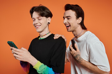 Two young friends, dressed in vibrant attire, pose against an orange backdrop, sharing a laugh while looking at a smartphone.