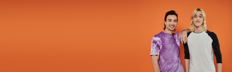 Two young queer friends in colorful clothing pose against an orange background.