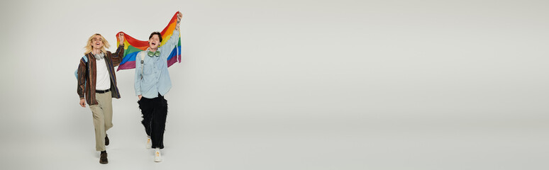 Obraz premium Two young students holding a rainbow pride flag and walking towards the camera on a grey background.