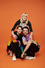 Fototapeta premium Three friends stand in front of a vibrant orange backdrop, wrapped in a pride flag, showing their LGBTQIA unity and solidarity.