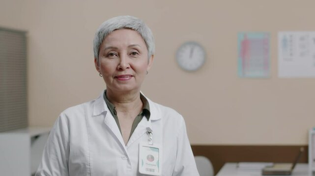 Portrait of mature Caucasian female gynecologist wearing white coat putting on badge and posing for camera in medical setting, getting ready for appointments