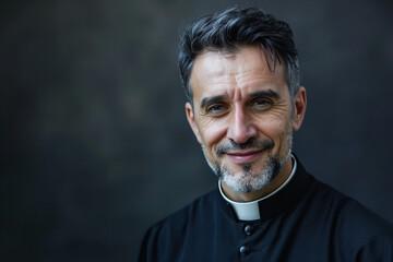 Fototapeta premium Good looking catholic priest is smiling on slack studio background. Religious man looks at the camera with serenity, peace and optimism. Copy paste empty place or text