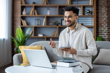 Obraz premium A young Indian male teacher teaches remotely, sits at home on the sofa with textbooks, makes notes in a notebook and talks on a video call