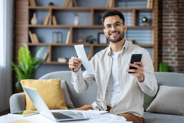 Portrait of a smiling and satisfied young man sitting at home on the sofa, holding a mobile phone and a message letter, looking happily at the camera