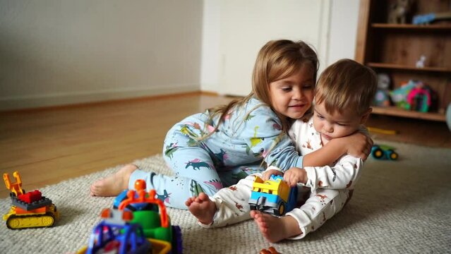 A sweet 7-month-old baby boy and his older sister sitting on the floor in a room, playing together with toys. The sister tenderly embraces her younger brother, capturing the theme of sibling love