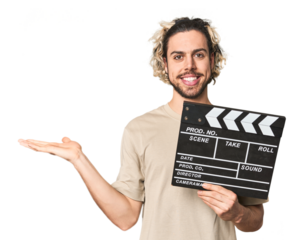 Young man with clapperboard in studio showing a copy space on a palm and holding another hand on waist.