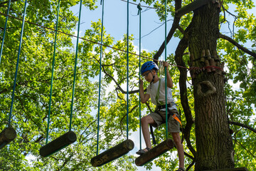 Obraz premium Boy exploring an outdoor playground with ropes and wooden obstacles. the child is wearing a helmet and safety gear, enjoying an adventure in nature on a sunny day. Rope trail in an adventure rope park