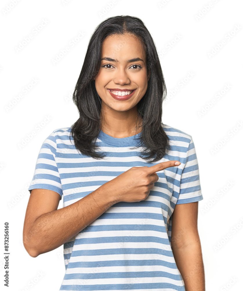 Wall mural young latina posing in studio smiling and pointing aside, showing something at blank space.