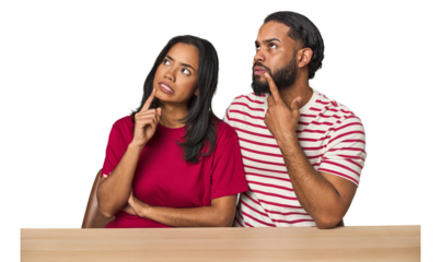 Seated young Latino couple at table looking sideways with doubtful and skeptical expression.