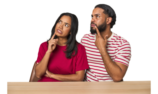 Seated young Latino couple at table looking sideways with doubtful and skeptical expression.