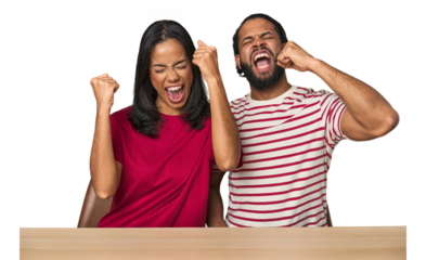 Seated young Latino couple at table raising fist after a victory, winner concept.