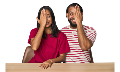 Seated young Latino couple at table having fun covering half of face with palm.