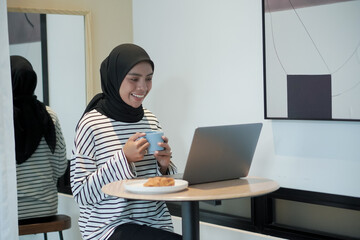 A woman wearing a black scarf and a white striped shirt is sitting at a table with a laptop and a cup of coffee. She is smiling and she is enjoying her coffee and laptop