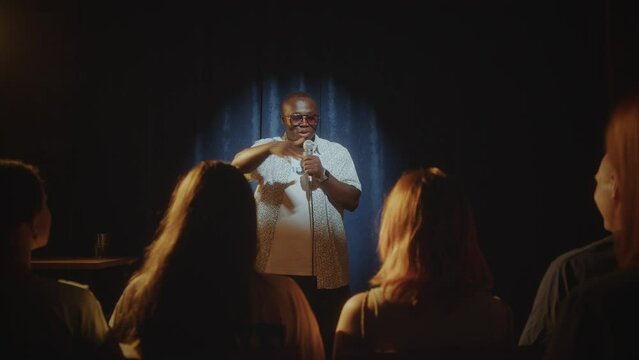 Young Black man standing on stage and talking in mic as performing stand-up comedy in front of audience. Zoom Shot