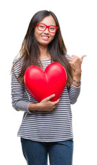 Young asian woman in love holding read heart over isolated background pointing and showing with thumb up to the side with happy face smiling