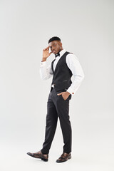 A young, handsome African American groom dressed in a black vest and tie, exuding elegance and charm in a studio shot on a grey background.